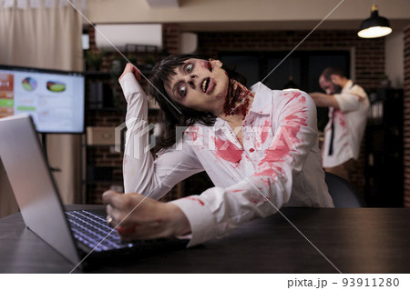 Horror cruel zombie using laptop at desk, undead corpse trying to work on computer in startup office. Creepy aggressive brain eating monster looking terrifying and horrible, sinister danger. 93911280