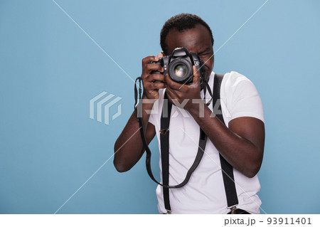 Confident professional photographer having DSLR camera taking picture. African american photography enthusiast taking photo while standing on blue background. Studio shot Confident professional photographer having DSLR camera taking picture. African american photography enthusiast taking photo while standing on blue background. Studio shot 93911401