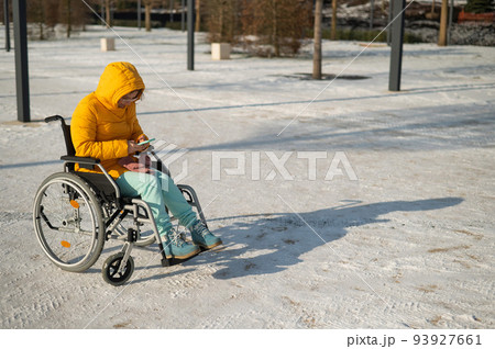 Caucasian woman in a wheelchair uses a smartphone on a walk in the park in winter.  93927661