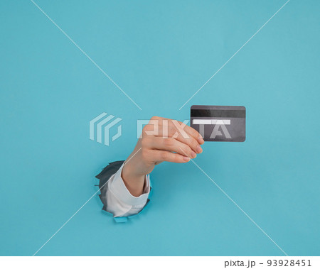 A woman's hand sticking out of a hole in a paper blue background holds a credit card. A woman's hand sticking out of a hole in a paper blue background holds a credit card. 93928451