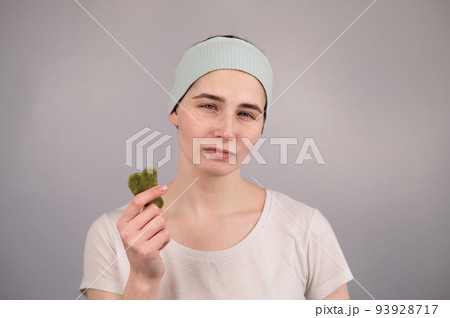 Portrait of a young woman massages her face with a gouache scraper on a white background. 93928717