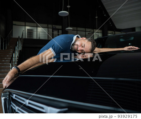 Happy caucasian man hugging the hood of his new car in a car dealership. 93929175