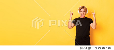 Sassy handsome asian guy likes heavy metal, listening hard rock music, showing rock-n-roll gesture and wearing wireless headphones, sticking tongue, standing yellow background Sassy handsome asian guy likes heavy metal, listening hard rock music, showing rock-n-roll gesture and wearing wireless headphones, sticking tongue, standing yellow background 93930736
