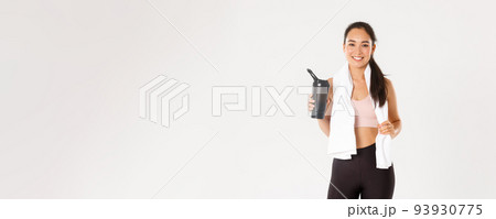 Sport, wellbeing and active lifestyle concept. Portrait of satisfied attractive asian fitness girl with cute smile, looking pleased wiping sweat with towel and drinking water after workout 93930775