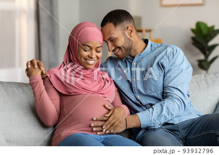 Married black muslim couple expecting baby cuddling on couch in living room Married black muslim couple expecting baby cuddling on couch in living room 93932796