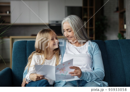 Smiling european small girl gives card to old woman, enjoy moment together in living room interior Smiling european small girl gives card to old woman, enjoy moment together in living room interior 93933058