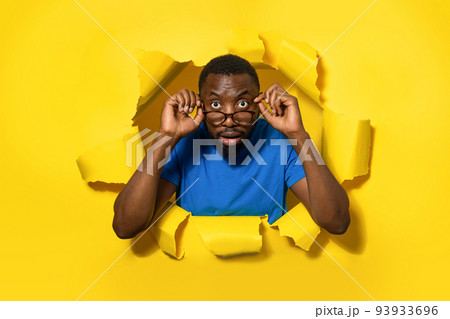 Emotional black man in eyeglasses looking at camera through hole in yellow paper, opening mouth with shock, banner 93933696