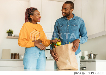 Ordering food online. Happy african american couple arriving from supermarket with grocery bag and unpacking in kitchen Ordering food online. Happy african american couple arriving from supermarket with grocery bag and unpacking in kitchen 93933699