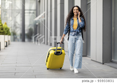 Smiling young woman traveller walking by airport, having phone conversation 93933831