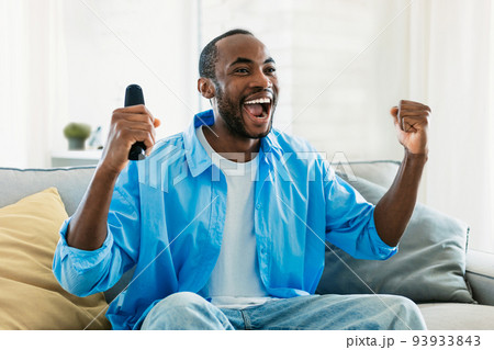 Overjoyed black man watching sport on TV at home, celebrating success, raising fists up and screaming, copy space Overjoyed black man watching sport on TV at home, celebrating success, raising fists up and screaming, copy space 93933843