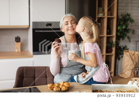 Cheerful european little granddaughter kisses her grandmother on cheek enjoy cookies with milk at kitchen Cheerful european little granddaughter kisses her grandmother on cheek enjoy cookies with milk at kitchen 93933912