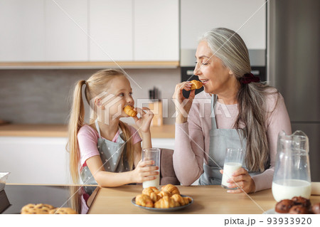 Cheerful caucasian aged grandmother and small granddaughter in aprons with glasses of milk, eat pastries 93933920