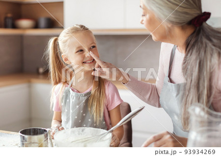 Happy senior woman in apron touches nose of little girl, make dough and have fun with flour, family enjoy leisure time Happy senior woman in apron touches nose of little girl, make dough and have fun with flour, family enjoy leisure time 93934269