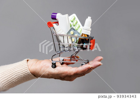 Hand of elderly woman rolls a full of medicines little shopping cart. Gray background. Concept of online shopping and sales 93934343