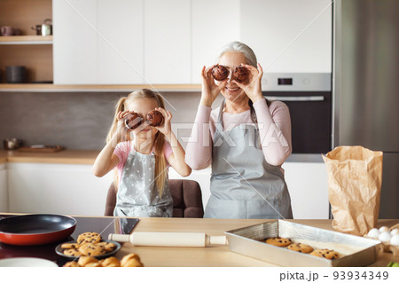 Happy european little granddaughter and old female have fun applying ready cookies to eyes Happy european little granddaughter and old female have fun applying ready cookies to eyes 93934349