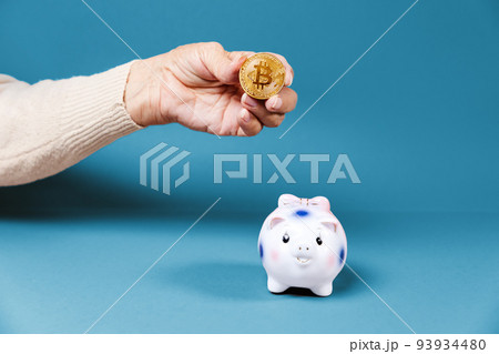 Wrinkled hand of an elderly woman show a bitcoin near a defocused piggy bank. Blue background. The concept of investment and cryptocurrency 93934480