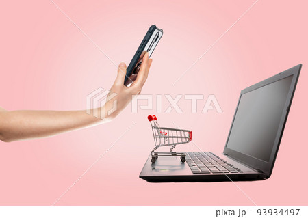 Modern technology. A laptop with a mini grocery cart standing on it, and a woman pays for a purchase with a smartphone. Pink background. The concept of online shopping Modern technology. A laptop with a mini grocery cart standing on it, and a woman pays for a purchase with a smartphone. Pink background. The concept of online shopping 93934497