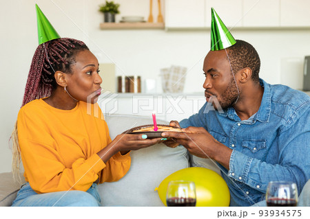 Happy black couple in party hats blowing lit candles on birthday pie, drinking wine and celebrating b-day together 93934575