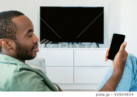 Black man watching television with blank screen and using smart TV remote control app on smartphone, mockup 93934654