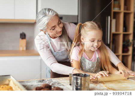 Cheerful caucasian elderly grandmother in apron teaching small girl to make dough with rolling pin for pizza and cookies Cheerful caucasian elderly grandmother in apron teaching small girl to make dough with rolling pin for pizza and cookies 93934718