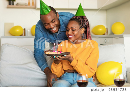 Happy black couple wearing party hats, celebrating birthday with festive cake at home, sitting on sofa and making wish 93934723