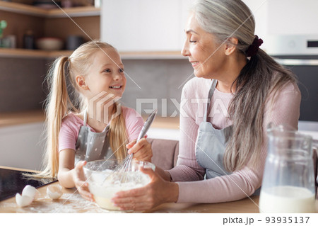 Happy european elderly grandmother and little granddaughter in aprons make pastry dough Happy european elderly grandmother and little granddaughter in aprons make pastry dough 93935137