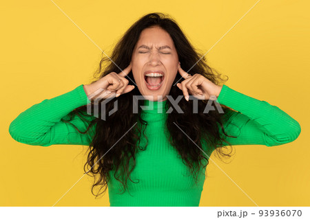 Woman Shouting With Eyes Closed Covering Ears Over Yellow Background 93936070