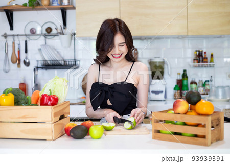 beautiful asian woman smiling preparing, cut green apple in white kitchen with plenty of fresh vegetables and fruits on the table. 93939391