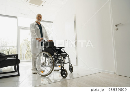 Professional senior male doctor holding wheelchair in hospital clinic hall 93939809