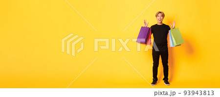 Full-length of happy handsome asian guy on shopping, holding bags and smiling, yellow background 93943813