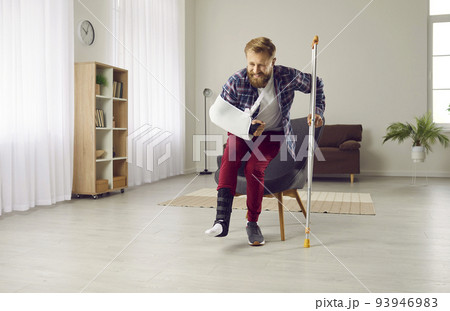 Man with broken leg and arm tries to get up from chair and walk with crutches at home. 93946983