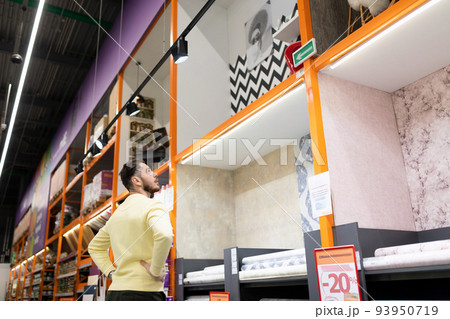 a man chooses finishing materials for repairs in his house next to the racks in a construction a man chooses finishing materials for repairs in his house next to the racks in a construction 93950719