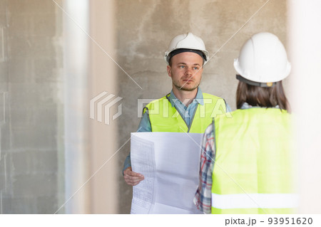 two engineers, a man and a woman in white helmets and protective vests, are standing in the room and holding a plan of the construction 93951620