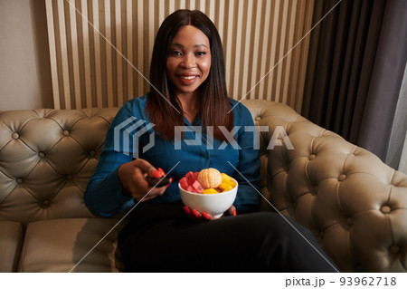Smiling Black woman sitting on couch with bowl of yeah fruits and turning on tv to watch new show Smiling Black woman sitting on couch with bowl of yeah fruits and turning on tv to watch new show 93962718