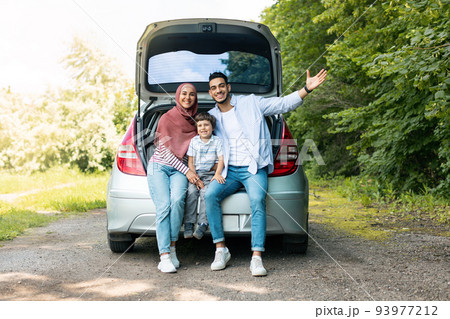 Positive auto journey. Happy millennial arabic guy and lady in hijab with baby, sit in car trunk and gesturing 93977212