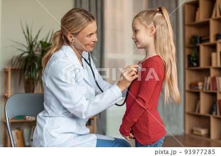 Cheerful female pediatrician listening girl's heartbeat with stethoscope during checkup at home, side view 93977285