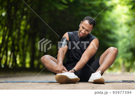 Handsome black sportsman exercising in public park, touching ankle Handsome black sportsman exercising in public park, touching ankle 93977394