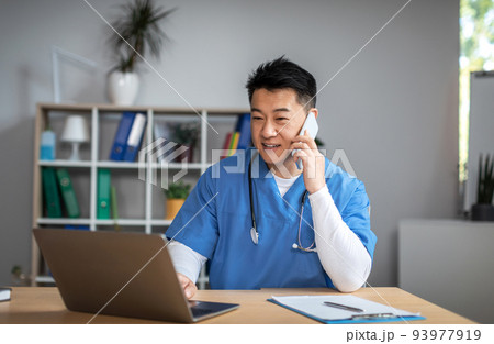 Smiling millennial asian man doctor in uniform call by phone, types on computer in office clinic interior Smiling millennial asian man doctor in uniform call by phone, types on computer in office clinic interior 93977919
