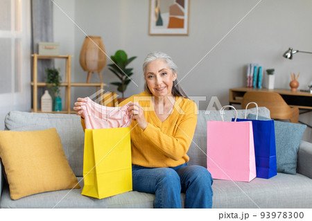 Cheerful caucasian senior gray-haired lady shows clothes from package, sits on sofa after shopping Cheerful caucasian senior gray-haired lady shows clothes from package, sits on sofa after shopping 93978300
