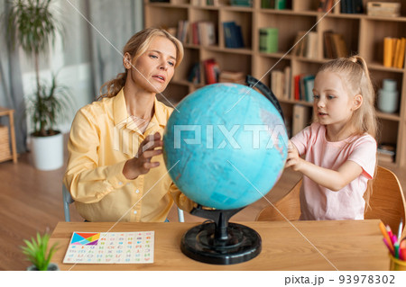 Professional preschool teacher teaching cute girl about globe, having private home class, sitting at desk in living room Professional preschool teacher teaching cute girl about globe, having private home class, sitting at desk in living room 93978302