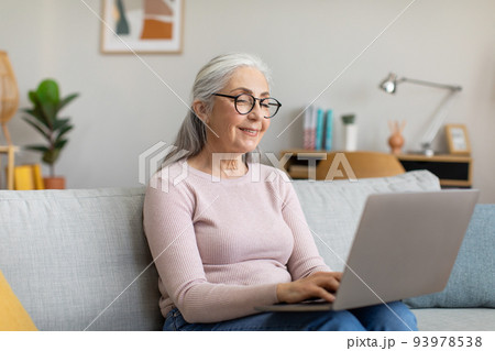 Happy caucasian mature gray-haired female in glasses typing on laptop, watching video in living room interior Happy caucasian mature gray-haired female in glasses typing on laptop, watching video in living room interior 93978538