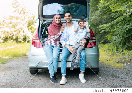 Smiling young middle eastern man and wife in hijab holding baby, sitting in car trunk, resting from trip outdoor Smiling young middle eastern man and wife in hijab holding baby, sitting in car trunk, resting from trip outdoor 93978600