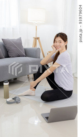 Beautiful Asian woman watching yoga teacher and light exercises in her living room at home, Watch live teaching or VDO on laptop, Dumbbells and a protein shake or bottle of water, Healthy exercise. 93979604