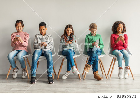 Multiracial Preteen Kids Using Cellphones Sitting Near Gray Wall Indoor Multiracial Preteen Kids Using Cellphones Sitting Near Gray Wall Indoor 93980871