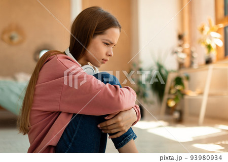 Teen depression concept. Stressed teenage girl sitting on floor at her bedroom, looking down, feeling upset and lonely Teen depression concept. Stressed teenage girl sitting on floor at her bedroom, looking down, feeling upset and lonely 93980934