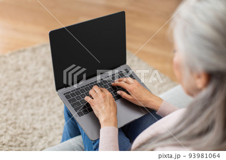 Cropped caucasian elderly gray-haired woman typing on laptop with blank screen in living room 93981064