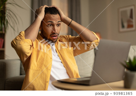 Shocked black guy looking at laptop screen, grabbing head in terror, making mistake, missing project deadline at home Shocked black guy looking at laptop screen, grabbing head in terror, making mistake, missing project deadline at home 93981365