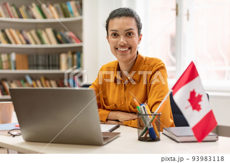 Online foreign languages tutoring. Happy female teacher sitting in library with flag of Canada, using laptop Online foreign languages tutoring. Happy female teacher sitting in library with flag of Canada, using laptop 93983118