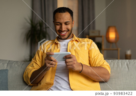 Happy young black man using smartphone, chatting online, sitting on couch at home Happy young black man using smartphone, chatting online, sitting on couch at home 93983422