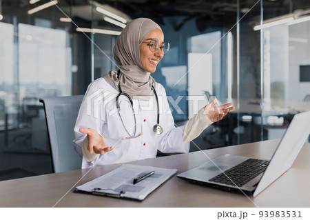 Doctor online consultation with patient, muslim woman in hijab working remotely with laptop for video call, consulting patients sitting inside modern clinic office Doctor online consultation with patient, muslim woman in hijab working remotely with laptop for video call, consulting patients sitting inside modern clinic office 93983531
